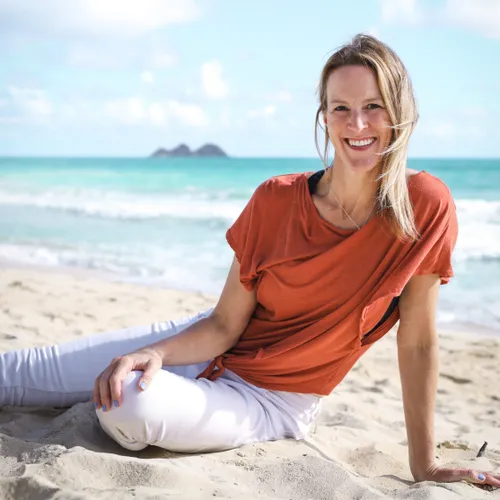 Smiling woman in an orange top and white pants sitting on sandy beach with ocean and blue sky in the background.